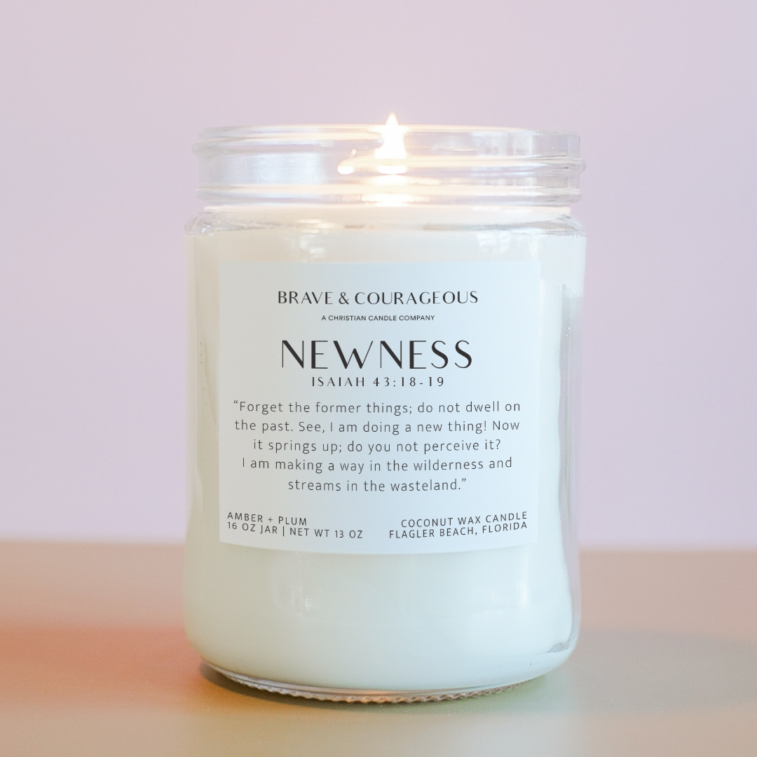 A white candle labeled 'NEWNESS' with the scent of amber and plum, featuring Bible verse from Isaiah 43:18-19, in a clear glass jar on a pink background.