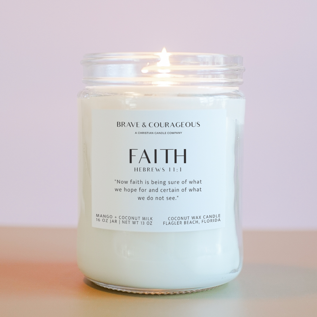 A lit candle in a clear glass jar with a white label that read "Brave & Courageous" and "Faith" with "Hebrews 11:1" on it in black text, on a light pink background.