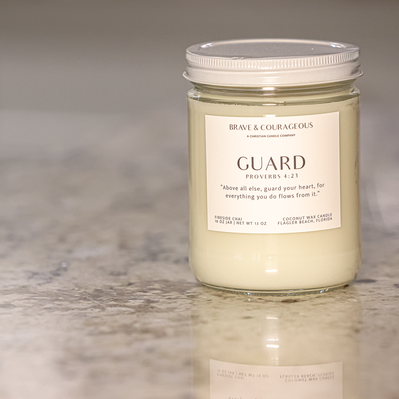 A glass jar candle with white wax and a white lid sits on a speckled granite countertop. The front label reads 'BRAVE & COURAGEOUS' and 'GUARD PROVERBs 4:23."