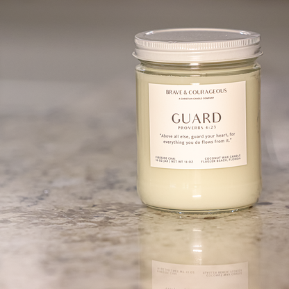 A glass jar candle with white wax and a white lid sits on a speckled granite countertop. The front label reads 'BRAVE & COURAGEOUS' and 'GUARD PROVERBs 4:23."