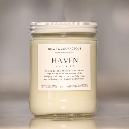 A glass jar candle with a white lid and a white label reading 'BRAVE & COURAGEOUS, HAVEN,  PSALM 91:1-2' on a neutral gray background.