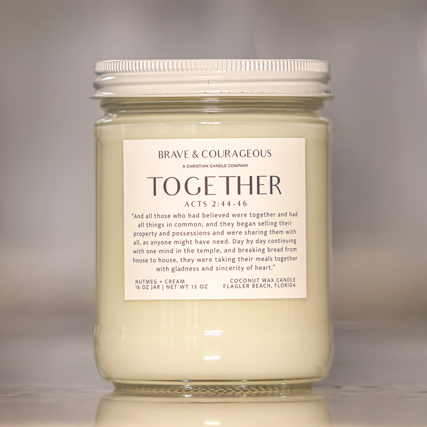 A glass jar candle with a white lid and a white label reading 'BRAVE & COURAGEOUS CANDLE COMPANY TOGETHER ACTS 2:44-46' on a reflective surface against a soft gray background.