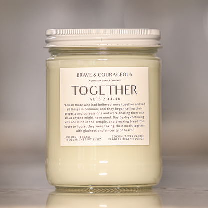 A glass jar candle with a white lid and a white label reading 'BRAVE & COURAGEOUS CANDLE COMPANY TOGETHER ACTS 2:44-46' on a reflective surface against a soft gray background.