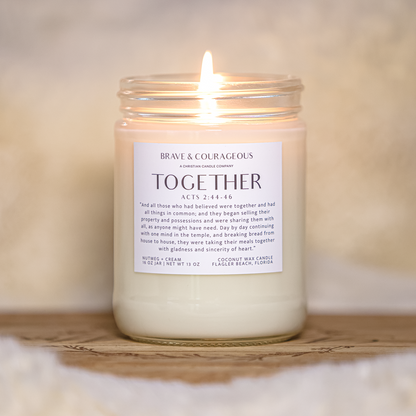 A lit candle in a clear glass jar sits on a wooden tray against a soft, warm neutral background. The white wax and front label read 'BRAVE & COURAGE ' and 'TOGETHER'.