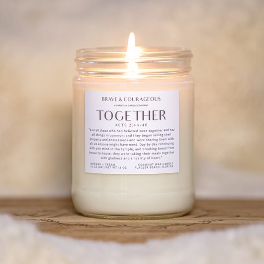 A lit candle in a clear glass jar sits on a wooden tray against a soft, warm neutral background. The white wax and front label read 'BRAVE & COURAGE ' and 'TOGETHER'.