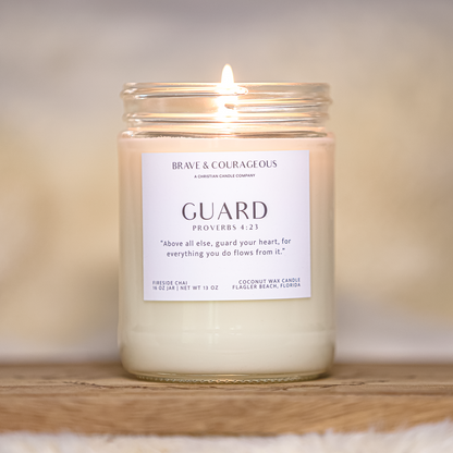 A lit coconut wax candle in a clear glass jar with a white label reading 'GUARD PROVERBS 4:23' and 'BRAVE & COURAGEOUS,' set on a wooden surface against a soft beige background.