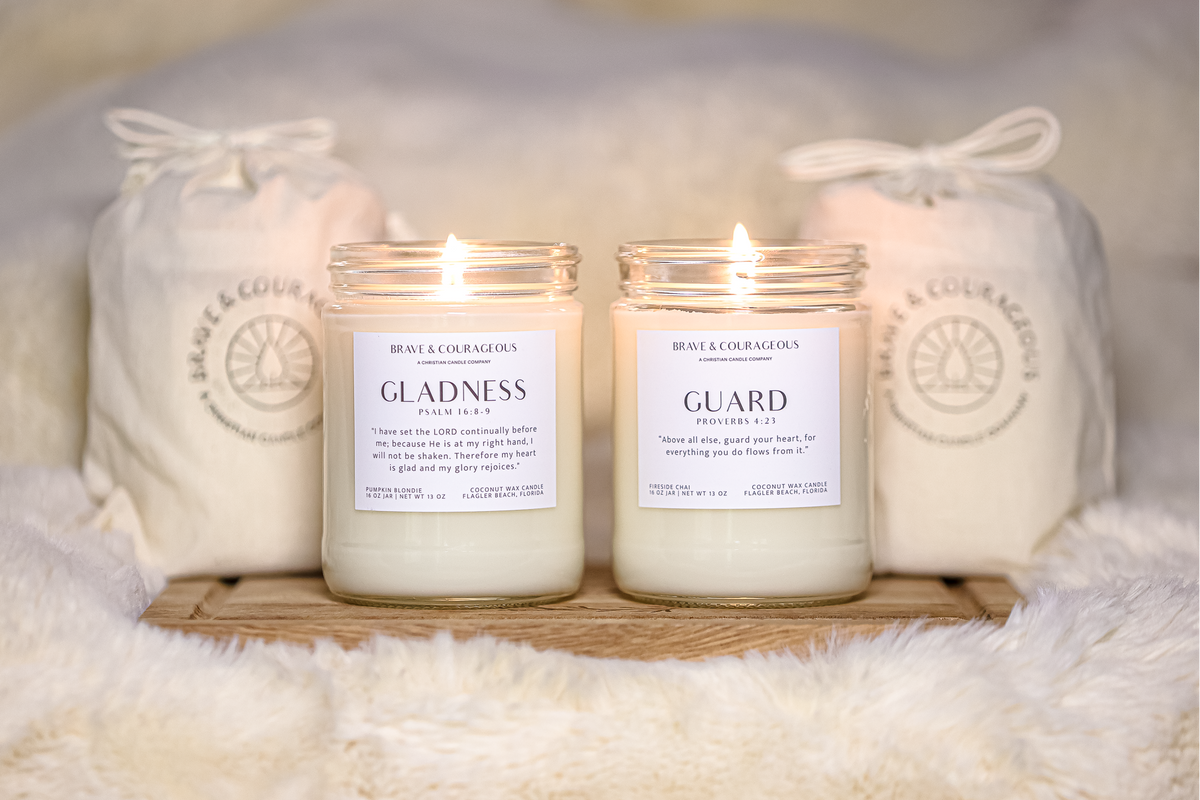 Two lit glass jar candles labeled "Gladness" and "Guard" by Brave & Courageous displayed on a wooden tray with two cream drawstring gift bags behind them on a soft white faux-fur surface.