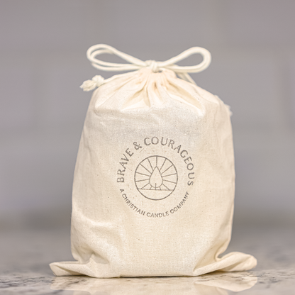 An off-white cotton drawstring bag with a printed circular logo reading 'BRAVE & COURAGEOUS' and 'A CHRISTIAN CANDLE COMPANY' on a marble countertop against a blurred light gray tiled background.