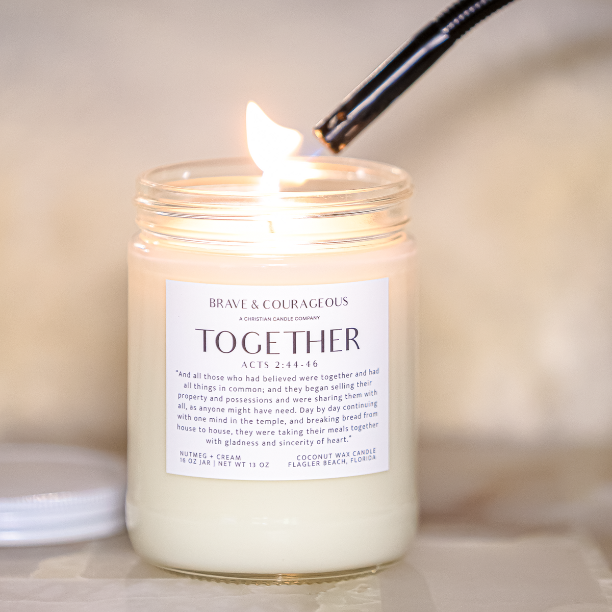 A lit candle in a clear glass jar with a white label reading 'BRAVE & COURAGEOUS Together' shown with a black lighter hovering above the flame. The lid rests beside the jar on a soft beige surface with a blurred warm background.