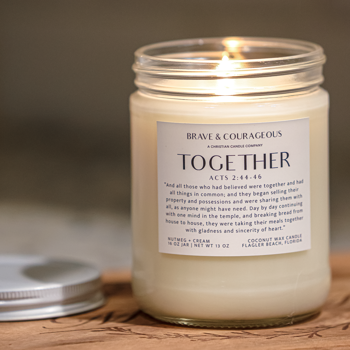 A lit coconut wax candle in a clear glass jar with a white label reading 'Brave & Courageous" "Together'  shown on a wooden surface with the metal lid beside it.