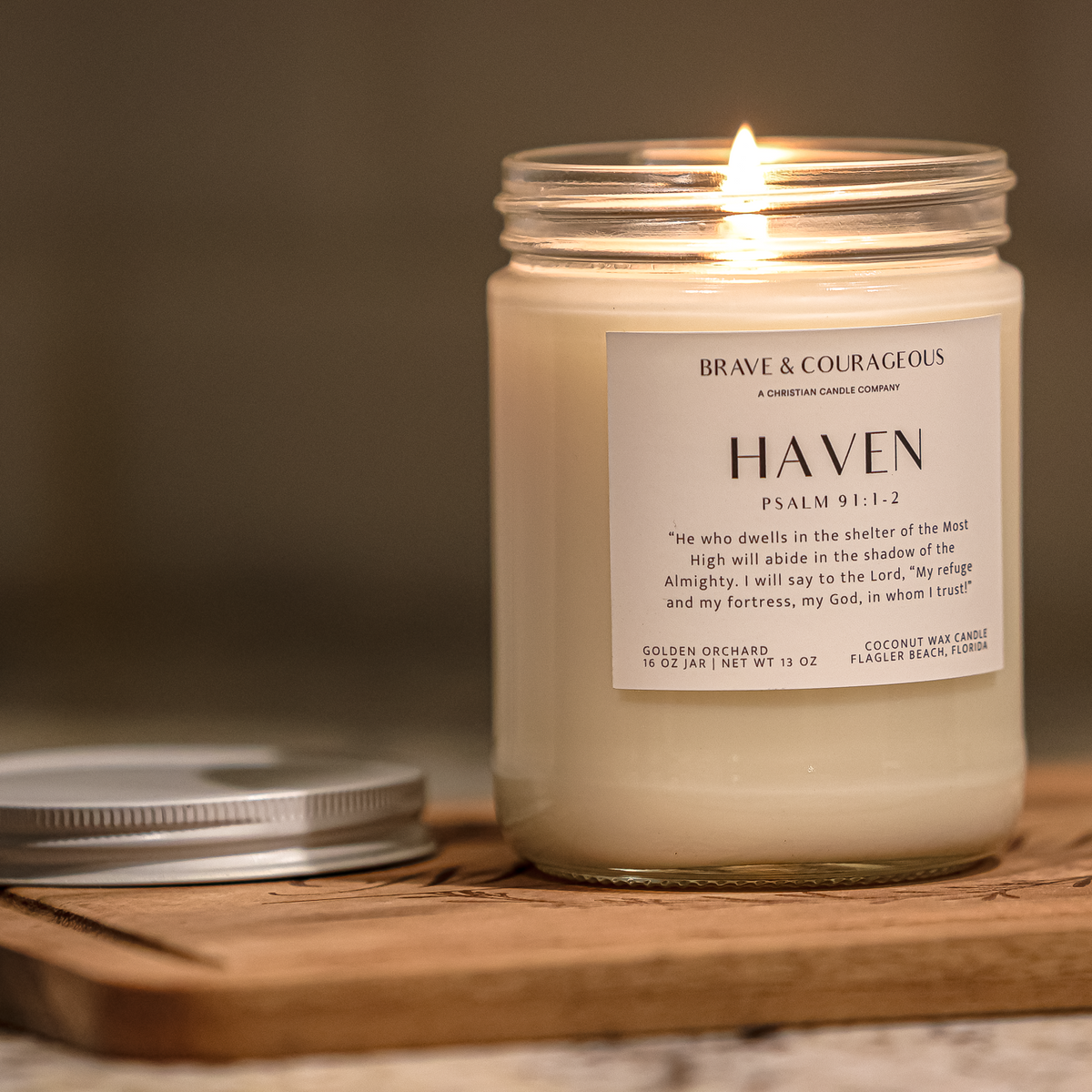 A lit glass jar candle labeled 'HAVEN' by Brave & Courageous, placed on a wooden board with its silver lid beside it in a warm indoor setting.