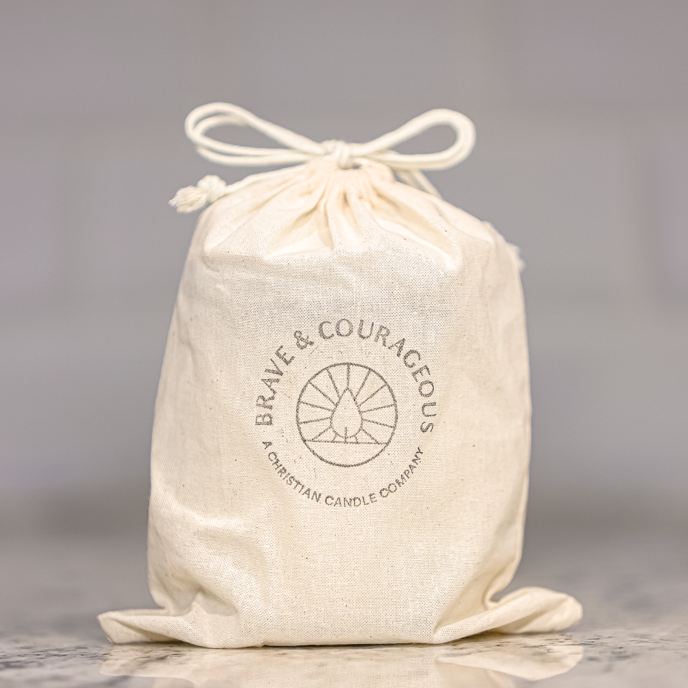 An off-white cotton drawstring bag with a printed circular logo reading 'BRAVE & COURAGEOUS' and 'A CHRISTIAN CANDLE COMPANY' on a marble countertop against a blurred light gray tiled background.