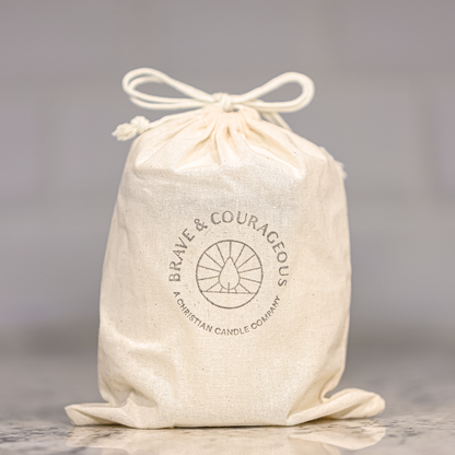 An off-white cotton drawstring bag with a printed circular logo reading 'BRAVE & COURAGEOUS' and 'A CHRISTIAN CANDLE COMPANY' on a marble countertop against a blurred light gray tiled background.