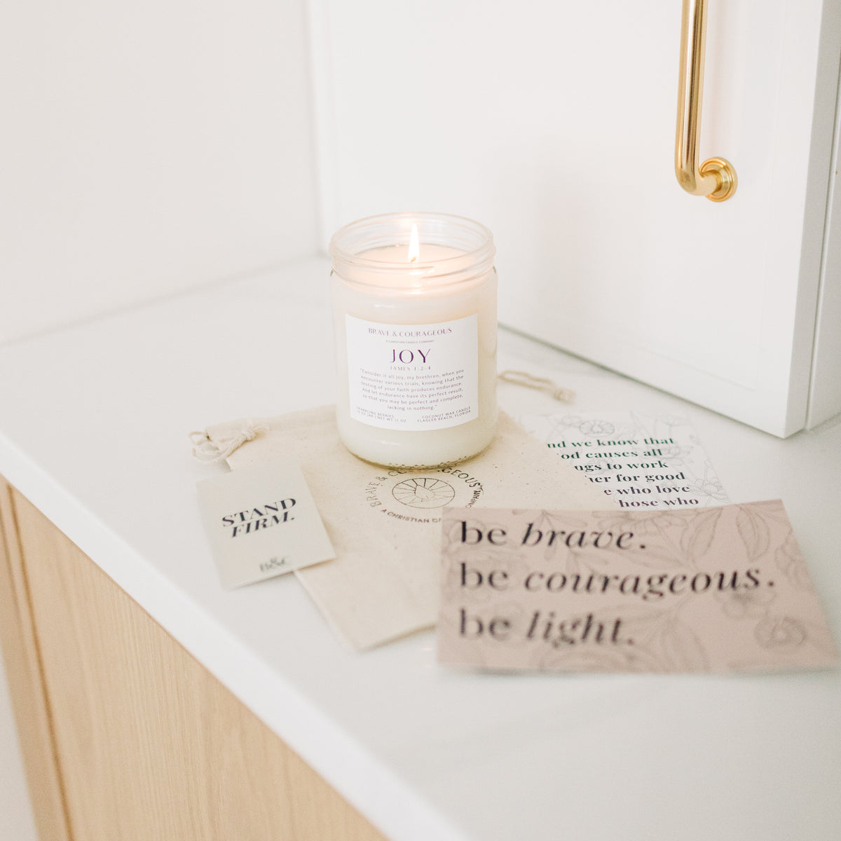 A lit white candle in a clear glass jar sits on a white countertop beside printed cards and a small tag. The candle label reads 'JOY' with James 1:2-4 underneath.