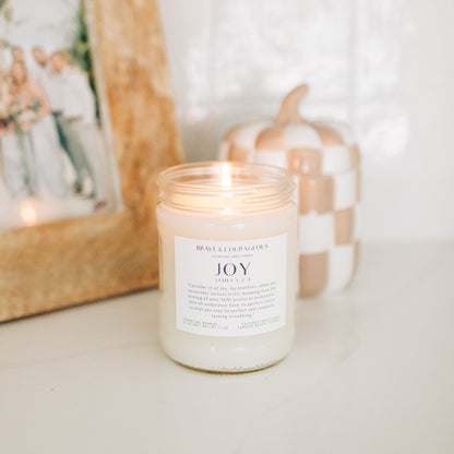 A lit white coconut wax candle in a clear glass jar with a white label reading 'Brave & Courage JOY', set on a light surface with a blurred framed photo and a checkered pumpkin in the background.
