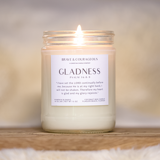 A lit glass jar candle with cream-colored wax sits on a wooden tray over a soft white faux-fur surface. The candle has a white label reading 'BRAVE & COURAGEOUS' and 'GLADNESS' with a Bible verse printed on it.