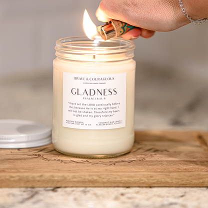 A glass jar candle labeled 'GLADNESS " is lit with a lighter on a wooden cutting board, with a silver lid resting beside it. A hand is visible holding the lighter, and the background is a softly blurred kitchen setting.