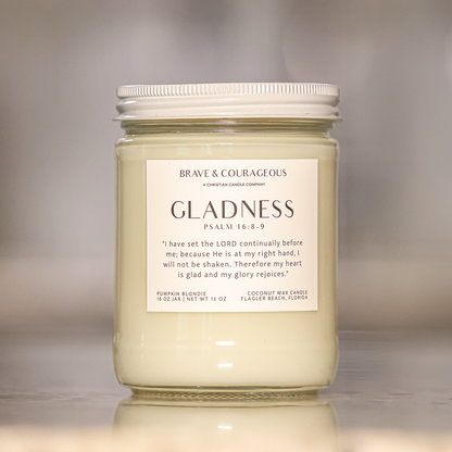 A jar candle with a white lid and a cream-colored wax body, featuring a white label that reads 'GLADNESS' and 'BRAVE & COURAGEOUS' with Bible verse Psalm 16:8-9. The candle is set on a reflective surface with a soft, blurred background.