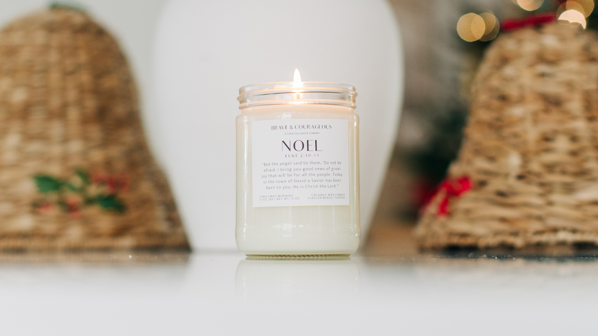A clear glass jar lit candle that reads Noel Brave and Courageous with a Bible verse underneath. It sits on a white counter top with some Christmas wicker bells and lights in the background.