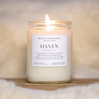 A lit glass jar candle with white wax sits on a wooden surface against a soft beige background. The front label reads 'Brave & COURAGEOUS' and 'HAVEN Psalm 91:1-2'.