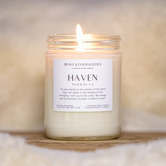 A lit glass jar candle with white wax sits on a wooden surface against a soft beige background. The front label reads 'Brave & COURAGEOUS' and 'HAVEN Psalm 91:1-2'.