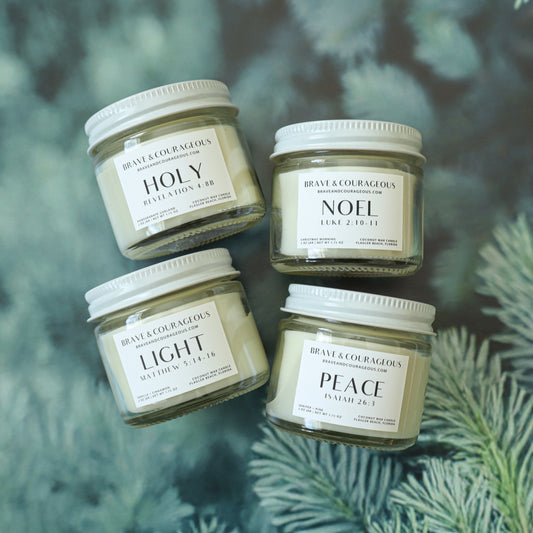 four white 2oz candles labeled "Holy" "noel" "light" and "peace" on a christmas tree background in flat lay design.