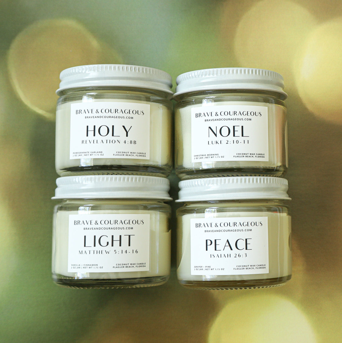 four 2oz clear candle jars with white labels that read "holy" "noel" "light" and "peace" with bible verse references on each. in a flat lay design with a gold colored background.