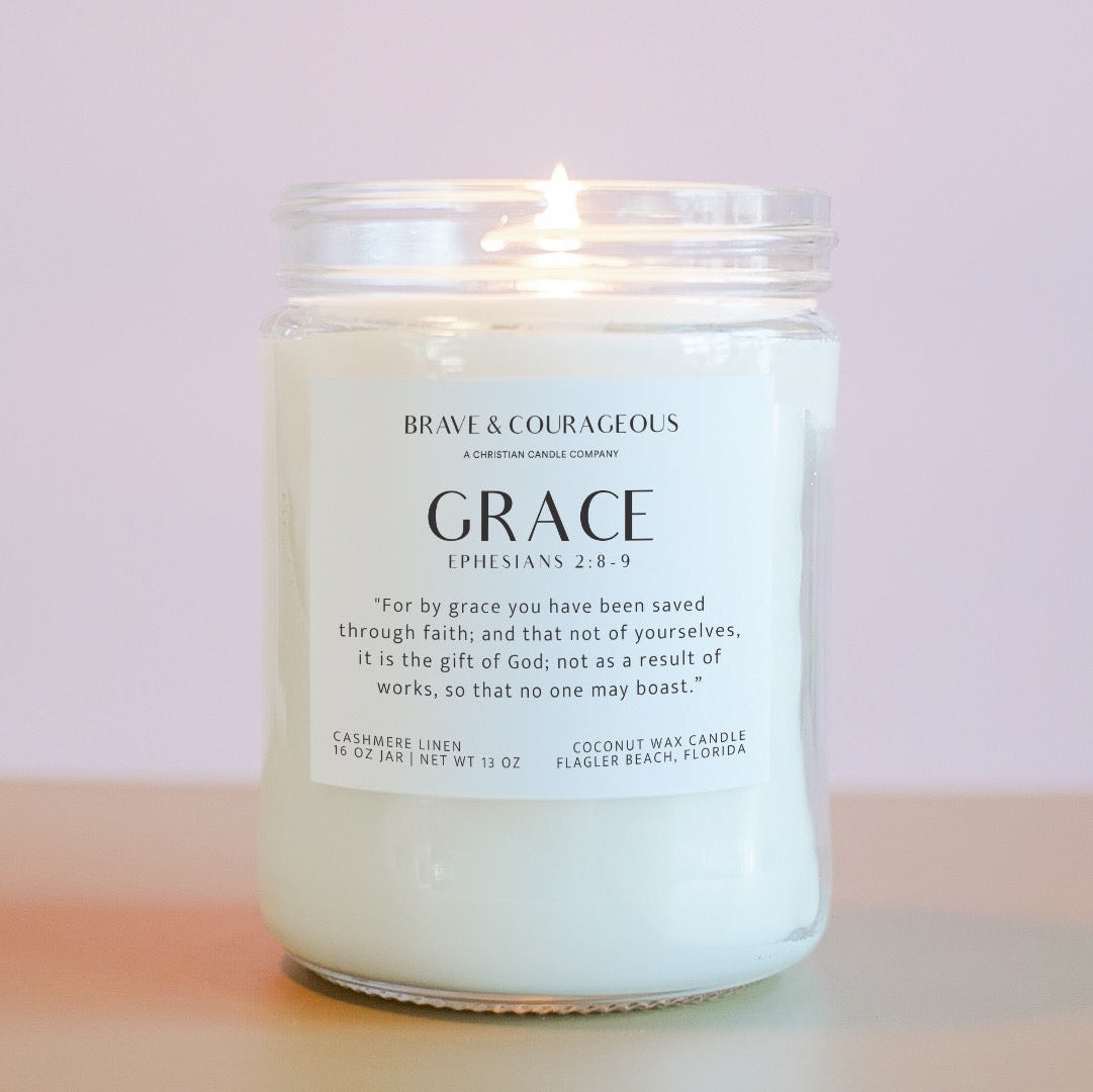 A lit candle in a clear glass jar with a white label that read "Brave & Courageous" and "Grace" with "Ephesians 6:8-9" on it in black text, on a light pink background.