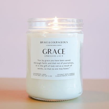 A lit candle in a clear glass jar with a white label that read "Brave & Courageous" and "Grace" with "Ephesians 6:8-9" on it in black text, on a light pink background.